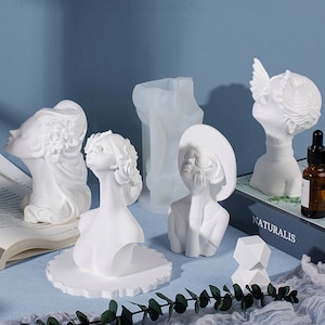 May include: Five white plaster sculptures of women's busts, one with wings, one wearing a hat and sunglasses, and two with floral headbands. The sculptures are arranged on a blue surface with a book and a small white cube.