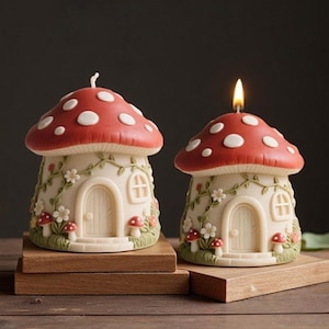 May include: Two mushroom-shaped candles. The candle on the right is lit. The mushroom caps are red with white spots. The candle bases are off-white with a door, windows, and floral accents. The candles sit on wooden blocks.