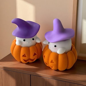 May include: Two Halloween-themed decorations. Each features a white ghost-like figure wearing a purple witch hat, nestled inside an orange pumpkin. The pumpkins have carved jack-o'-lantern faces. The decorations are on a wooden surface.