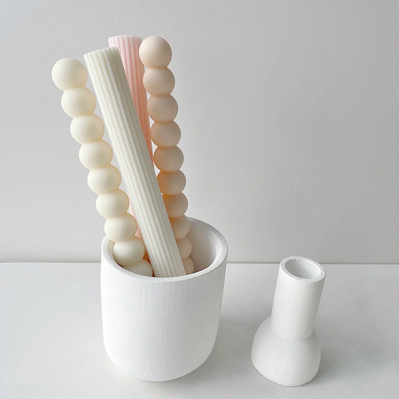 May include: Three decorative candles in a white ceramic holder. The candles are in different shapes and colors: one is a beaded white candle, one is a ribbed white candle, and one is a smooth pink candle.