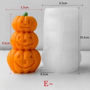 K&ouml;nnte beinhalten: Eine orangefarbene, gestapelte K&uuml;rbisskulptur mit geschnitzten Jack-o'-Lantern-Gesichtern, 9,5 cm hoch und 5,5 cm breit. Daneben eine wei&szlig;e, zylindrische Form, 10,5 cm hoch und 6,5 cm breit. Der Buchstabe "E~" befindet sich unten.