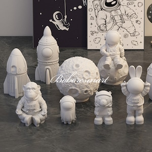 May include: A set of white astronaut figurines, including a monkey, a bear, a rabbit, and a person. The figurines are sitting on a moon, a rocket, and a spaceship. The figurines are made of a smooth, white material.