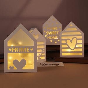 May include: White wooden house-shaped lanterns with cut-out designs, including hearts and the word "HOME." The lanterns are illuminated with warm, soft lighting, creating a cozy ambiance. The phrase "HOME SWEET HOME" is also visible on one of the lanterns.