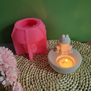 May include: A pink silicone mold shaped like a mushroom, and a white ceramic candle holder with a frog figurine on top. The candle holder has a small candle inside.