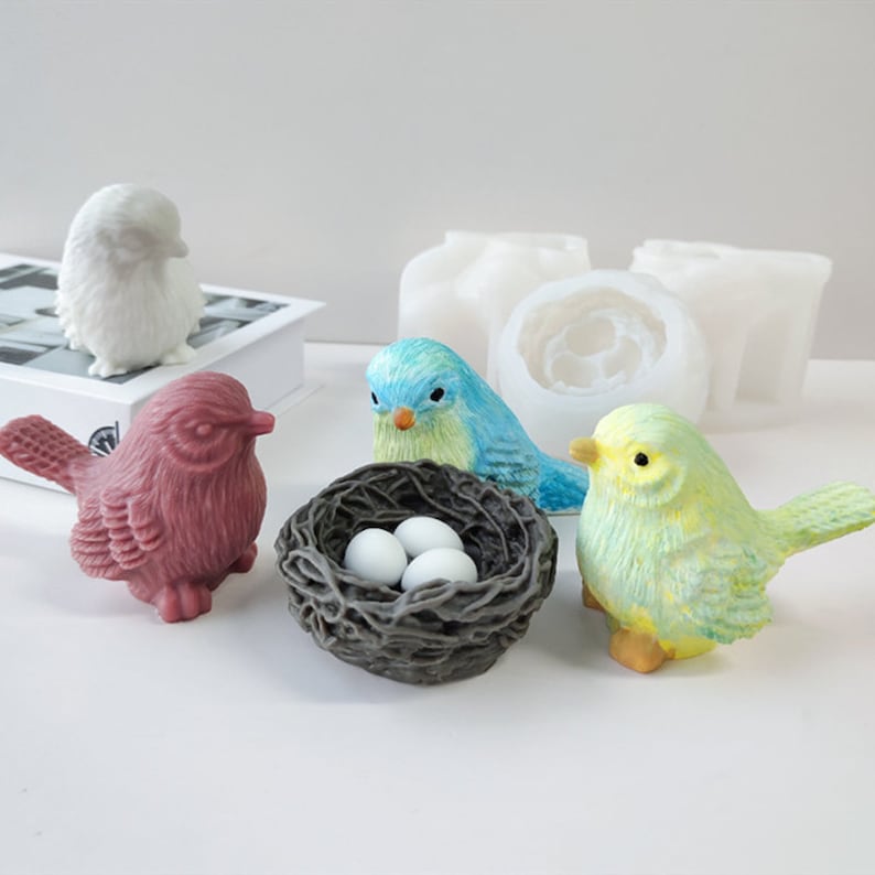 May include: A selection of colourful bird figurines and a nest with eggs. The birds are in shades of white, pink, blue, and yellow. A white book and several white moulds are also in the image.