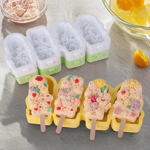 Może przedstawiać: Żółta silikonowa forma z czterema formami na lody na patyku z kwiatowymi wzorami. Cztery lody na patyku z kwiatowymi wzorami znajdują się w formie. Lody na patyku mają drewniane patyczki.