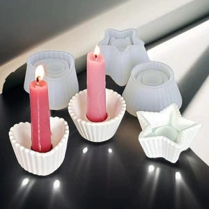 Puede incluir: Seis portavelas blancos de diferentes formas, incluyendo una estrella, un círculo y una flor. Dos velas rosas están en los portavelas.