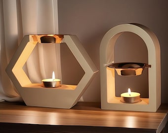 May include: Two beige ceramic candle holders with a gold metal top. One holder is shaped like a hexagon and the other is shaped like an arch. Both holders have a small candle inside.