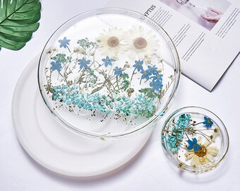 May include: Clear resin coasters with embedded dried flowers. The coasters feature white daisies, blue forget-me-nots, and other greenery. A larger coaster sits atop a white, round base, with a smaller coaster to the side.