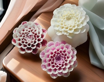 May include: Three decorative wax candles shaped like white and pink dahlias. The candles are on a wooden surface.