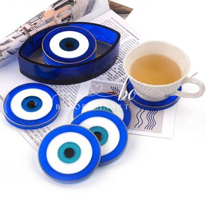 Op de afbeelding: Set van zes blauwe en witte boze oog-onderzetters met gouden rand. De onderzetters zijn rond en hebben een blauw oog met een witte pupil en een gouden ring rond het oog. De onderzetters zijn in een cirkel gerangschikt rond een grotere, ovale boze oog-onderzetter.
