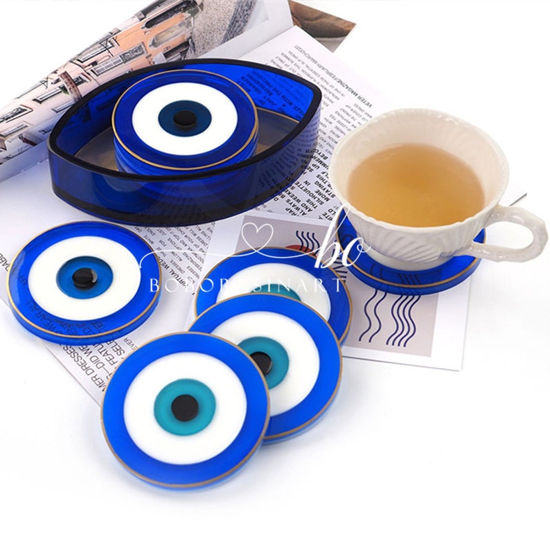 Op de afbeelding: Set van zes blauwe en witte boze oog-onderzetters met gouden rand. De onderzetters zijn rond en hebben een blauw oog met een witte pupil en een gouden ring rond het oog. De onderzetters zijn in een cirkel gerangschikt rond een grotere, ovale boze oog-onderzetter.