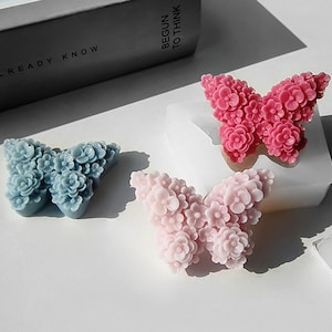 Peut inclure: Trois décorations en forme de papillon, de couleurs bleu clair et rose. Chaque papillon est composé de petites fleurs. Les objets sont présentés sur une surface blanche. Mesures en centimètres.