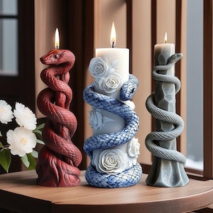 Puede incluir: Tres velas de columna decorativas. Una presenta un diseño de serpiente roja, otra una serpiente azul y motivos florales, y la tercera una serpiente gris. Todas tienen mechas encendidas y están sobre una superficie de madera.