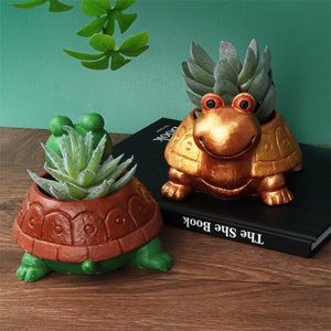 May include: Two turtle-shaped planters, one gold and one brown, with green legs and heads. Each planter has a succulent plant inside.