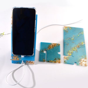 K&ouml;nnte beinhalten: Ein blauer und goldener Telefonst&auml;nder aus Harz mit einem Ladeanschluss und einem passenden kleineren St&auml;nder. Der Telefonst&auml;nder ist so konzipiert, dass er ein Telefon aufrecht h&auml;lt.