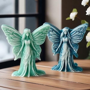 Op de afbeelding: Twee feeënbeeldjes, één in teal en de andere in blauw, staan op een houten oppervlak. Elke fee heeft gedetailleerde vleugels en vloeiende jurken. De beeldjes zijn decoratief en artistiek.