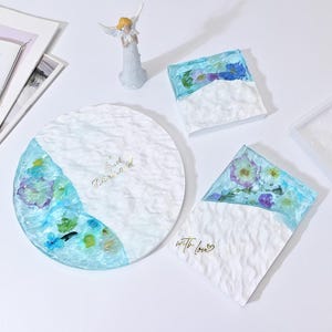 Könnte beinhalten: Drei Harztabletts mit einer weißen, strukturierten Oberfläche und einem blauen Rand. Die Tabletts zeigen gepresste Blumen und den Text "Soul Divine" und "With Love".