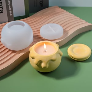 Puede incluir: Una vela amarilla en un diseño de cera que gotea, con una mecha blanca y una llama encendida. La vela está en un molde de silicona, con una tapa separada. El molde está sobre una superficie de madera.