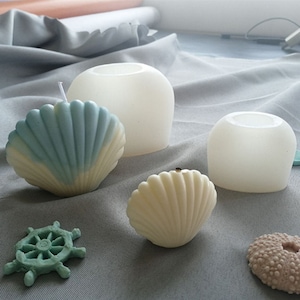 May include: Two white silicone moulds shaped like seashells, one with a blue and white seashell candle inside, and another with a white seashell candle inside. There is also a white silicone mould shaped like a round pillar candle and a blue and green seashell-shaped candle holder.