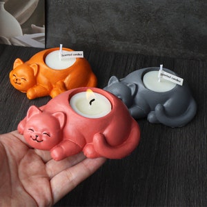 Può includere: Tre portacandele in ceramica a forma di gatto in arancione, rosso e grigio. Ogni portacandele presenta un design di gatto che dorme e un piccolo spazio rotondo per una candela tealight. Il testo "Candele profumate" è scritto su un piccolo cartellino bianco attaccato a ogni portacandele.