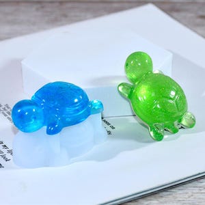 May include: Two small turtle figurines made of clear resin with glitter. The turtle on the left is blue and the turtle on the right is green.