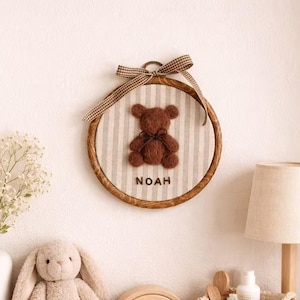May include: A brown teddy bear applique on a striped fabric, framed in a wooden hoop. The name "NOAH" is below the bear. A decorative ribbon is tied at the top. The scene includes a stuffed bunny, a wooden rainbow, and a lamp.