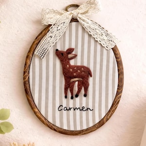 Op de afbeelding: Een ovale wanddecoratie in een borduurring met een bruine vilt-hertenapplicatie op een beige en wit gestreepte stoffen achtergrond. De naam Carmen is in zwarte draad onder het hert geborduurd, met een witte kanten strik aan de bovenkant.