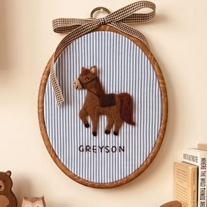 May include: An oval wooden embroidery hoop frame containing blue and white pinstriped fabric. A brown felt horse appliqué is centred above the embroidered name GREYSON. A brown and white gingham ribbon is tied in a bow at the top.