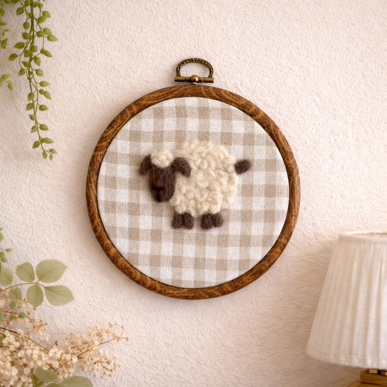 Peut inclure: D&eacute;coration murale artisanale avec un motif de mouton. Le mouton est fait de fil textur&eacute; blanc cass&eacute; avec des accents bruns, sur un tissu vichy beige et blanc. L'&oelig;uvre est encadr&eacute;e dans un cercle en bois avec une boucle en m&eacute;tal pour l'accrocher.