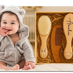 Puede incluir: Un juego de regalo para bebés con un cepillo, un peine y un cepillo de madera con cerdas suaves, presentado en una caja de regalo. El cepillo y el peine tienen ilustraciones de animales. El bebé lleva una chaqueta con capucha gris.