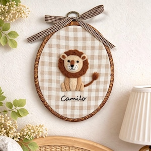 Peut inclure: Une décoration murale ovale sur tambour à broder avec un fond en tissu vichy beige et blanc. Une applique en feutrine représentant un lion est placée au centre au-dessus du nom brodé Camilo. Un ruban à carreaux marron et blanc est noué en haut.