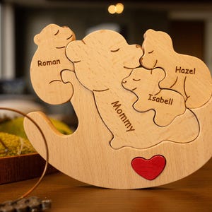 Può includere: Puzzle in legno con cinque pezzi a forma di orso, ognuno con un nome: Mommy, Roman, Hazel e Isabell. Il puzzle ha la forma di un orso e ha un cuore rosso al centro.