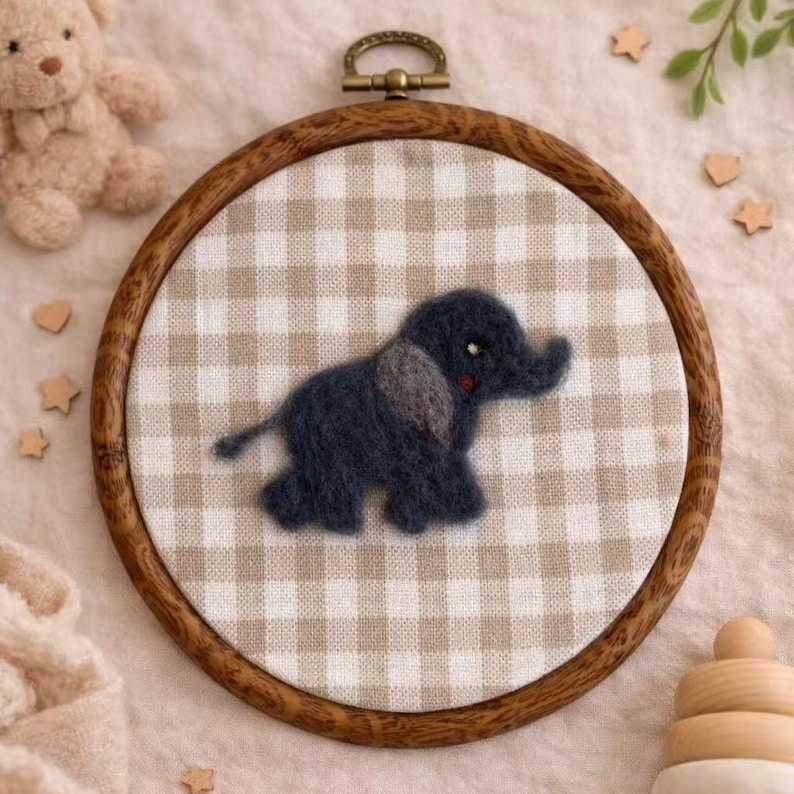 Peut inclure: Broderie artisanale d'un &eacute;l&eacute;phant en feutre dans un cercle en bois. L'&eacute;l&eacute;phant bleu fonc&eacute; avec des oreilles grises et des joues rouges est pos&eacute; sur un tissu vichy beige et blanc. Le cercle en bois a une boucle de suspension en m&eacute;tal. &Eacute;toiles en bois et ours en peluche en arri&egrave;re-plan.