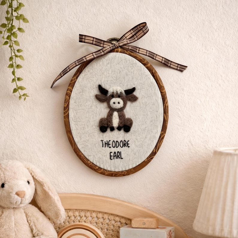 Peut inclure: D&eacute;coration murale ovale avec un cadre en bois marron et un motif de taureau en feutre. Le taureau est marron, blanc et noir. Le texte "THEODORE EARL" est en dessous. Un ruban &agrave; carreaux marron et beige est attach&eacute; en haut.