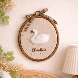 Peut inclure: D&eacute;coration murale ronde avec un cadre en bois, repr&eacute;sentant un cygne blanc sur un fond beige. Le nom "Charlotte" est brod&eacute; en dessous. Un n&oelig;ud en ruban &agrave; carreaux marron et blanc orne le haut.