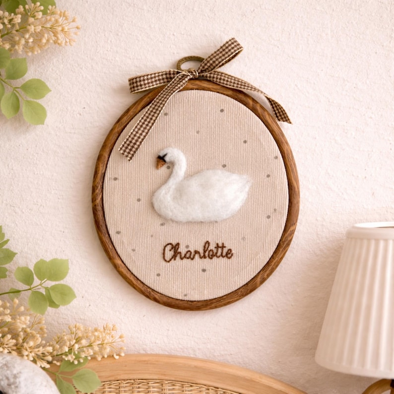Peut inclure: D&eacute;coration murale ronde avec un cadre en bois, repr&eacute;sentant un cygne blanc sur un fond beige. Le nom "Charlotte" est brod&eacute; en dessous. Un n&oelig;ud en ruban &agrave; carreaux marron et blanc orne le haut.