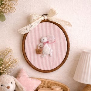 Peut inclure: D&eacute;coration murale artisanale sur le th&egrave;me de P&acirc;ques. Un lapin blanc en feutre avec un n&oelig;ud rose porte un panier, sur un fond &agrave; rayures roses et blanches. L'&oelig;uvre est encadr&eacute;e dans un cercle en bois et orn&eacute;e d'un n&oelig;ud en dentelle. Un charmant &eacute;l&eacute;ment de d&eacute;coration int&eacute;rieure.
