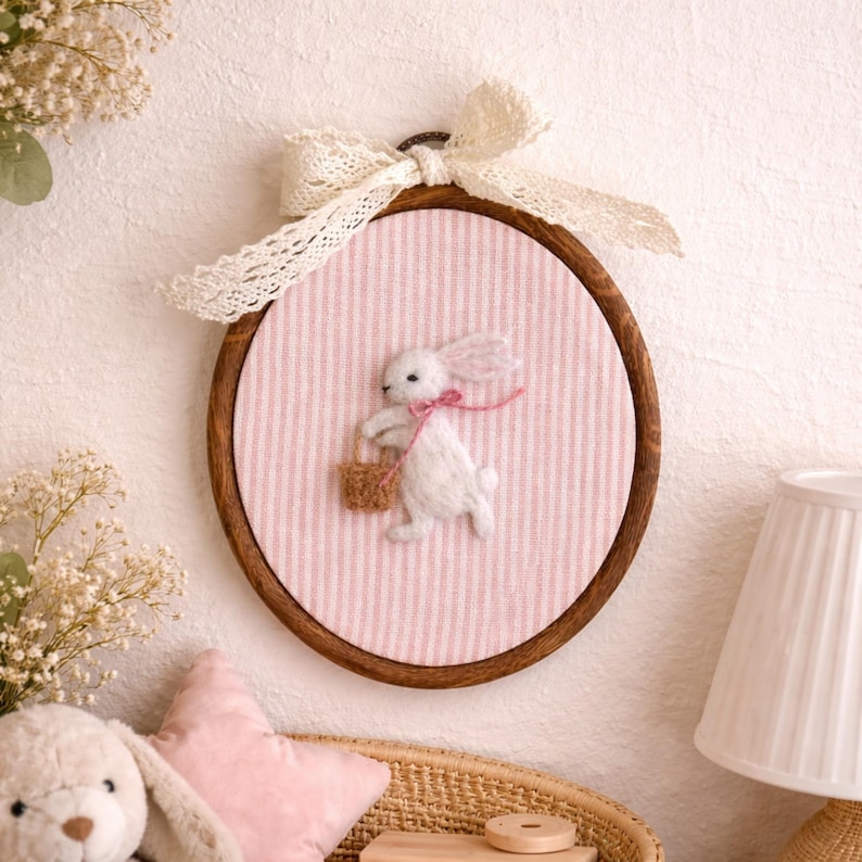Peut inclure: D&eacute;coration murale artisanale sur le th&egrave;me de P&acirc;ques. Un lapin blanc en feutre avec un n&oelig;ud rose porte un panier, sur un fond &agrave; rayures roses et blanches. L'&oelig;uvre est encadr&eacute;e dans un cercle en bois et orn&eacute;e d'un n&oelig;ud en dentelle. Un charmant &eacute;l&eacute;ment de d&eacute;coration int&eacute;rieure.