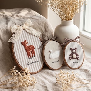 May include: Three oval wooden embroidery hoops featuring needle-felted designs: a brown deer labelled Carmen, a white swan labelled HARLIE, and a brown teddy bear labelled JACOB. Each hoop is decorated with a fabric bow and striped or solid fabric.