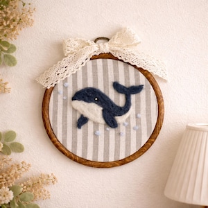Peut inclure: Broderie artisanale d'une baleine dans un cercle en bois. La baleine est bleu marine et blanche, sur un fond &agrave; rayures grises et blanches. Le cercle est orn&eacute; d'un n&oelig;ud en dentelle cr&egrave;me et de petits embellissements bleus.