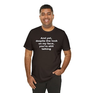 以下が含まれることがあります： 「And yet, despite the look on my face, you're still talking.」という白い文字が書かれた黒いTシャツです。
