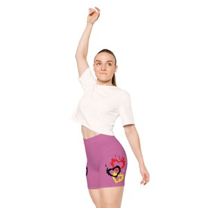 May include: A woman wearing a white t-shirt and pink athletic shorts with a black, red, and yellow heart design.