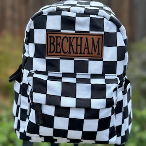 May include: A black and white chequered backpack with a front pocket and side pockets. The backpack features a brown leather patch with the name "BECKHAM" in capital letters.