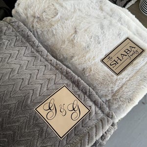 May include: Two plush blankets, one light gray with a chevron pattern and the other off-white with a textured surface. Each blanket has a wooden patch with custom text: "G & G" and "The SHABA Family".