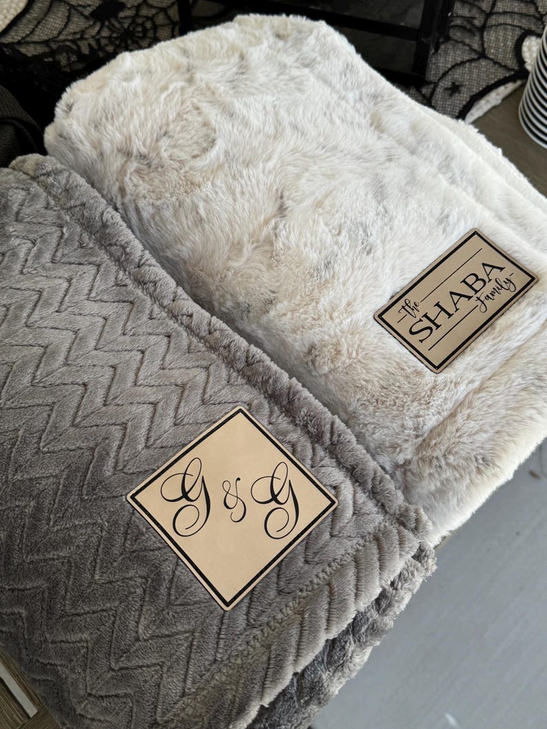 May include: Two plush blankets, one light gray with a chevron pattern and the other off-white with a textured surface. Each blanket has a wooden patch with custom text: "G & G" and "The SHABA Family".