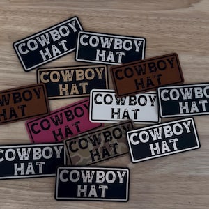 May include: A selection of rectangular patches, each bearing the text "COWBOY HAT" in a bold font. The patches are in various colors, such as black, brown, white, pink, and camouflage. These patches are likely designed for clothing or accessories.