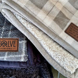 May include: Two plush blankets with a plaid pattern in gray and beige. One blanket has a leather patch with the text "Evolve Exteriors". The other blanket has a leather patch with the text "No matter the day, no matter the weather, life is just better when we are together."
