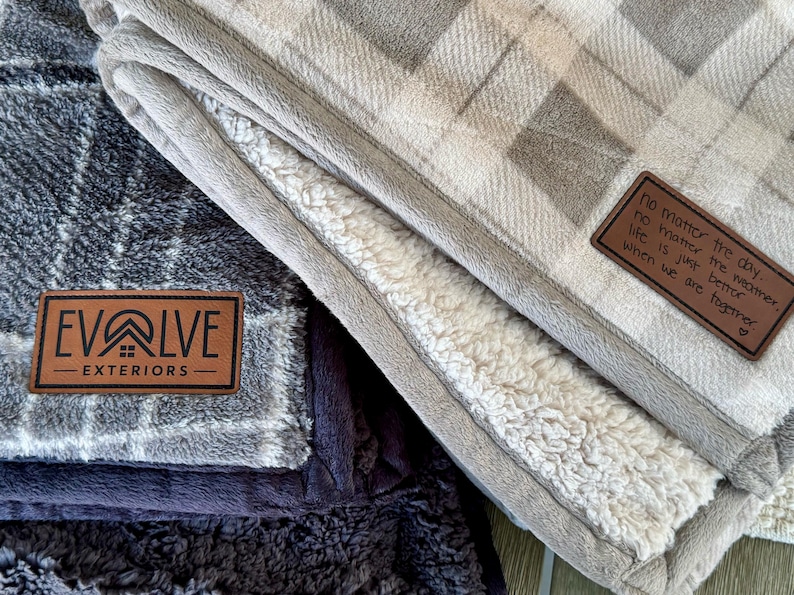 May include: Two plush blankets with a plaid pattern in gray and beige. One blanket has a leather patch with the text "Evolve Exteriors". The other blanket has a leather patch with the text "No matter the day, no matter the weather, life is just better when we are together."