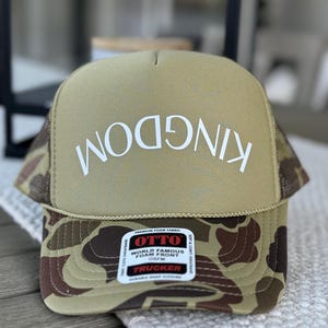 May include: A khaki trucker hat with a camouflage brim. The front of the hat has the text "KINGDOM" written in white.